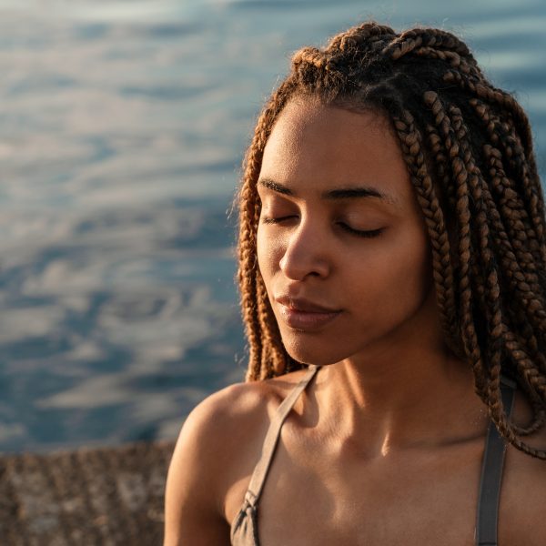 A person with braided hair sits by the water, embodying mindfulness in their daily life, eyes closed in serene calmness.