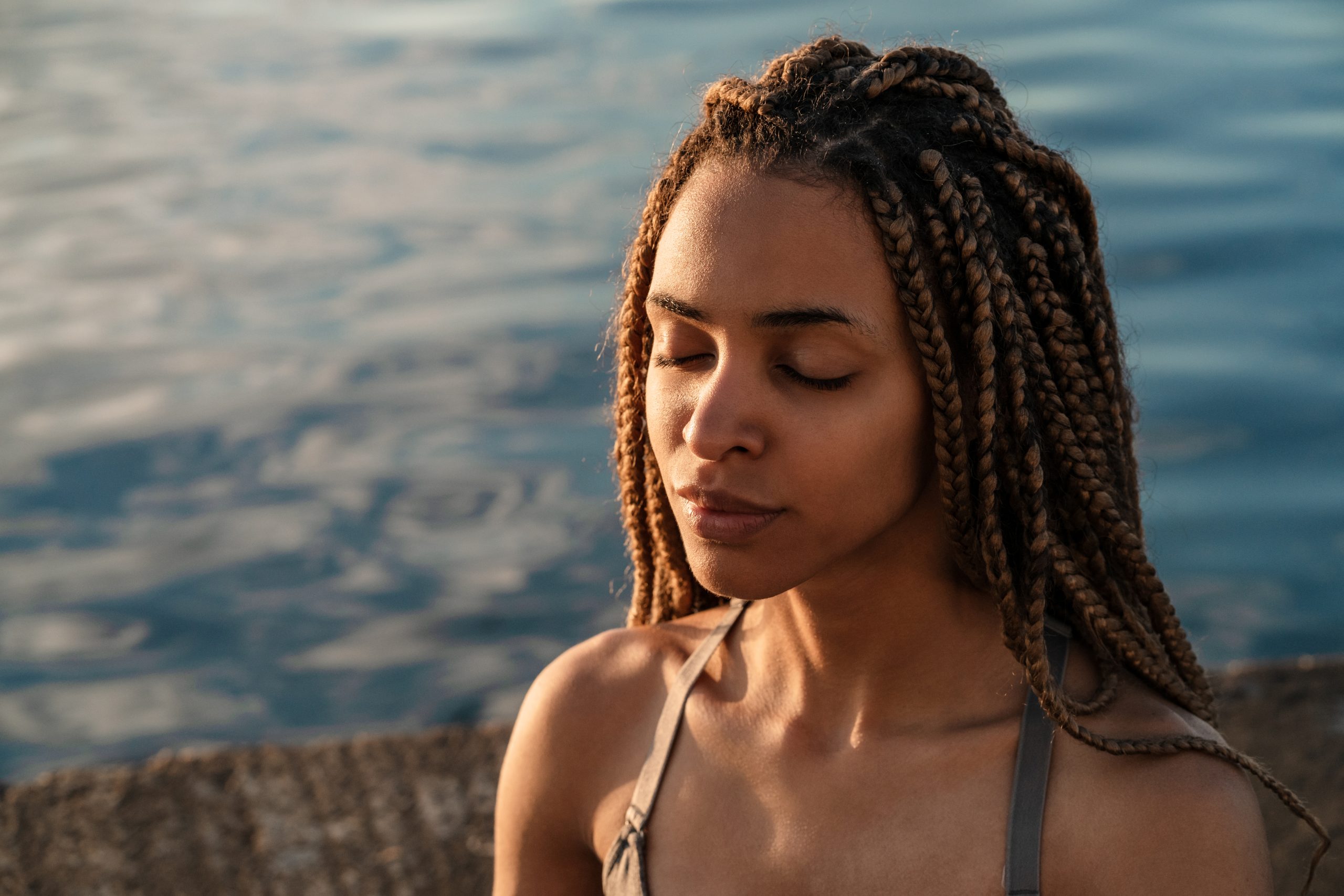 A person with braided hair sits by the water, embodying mindfulness in their daily life, eyes closed in serene calmness.