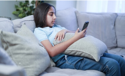 Young person sitting on a couch, holding a smartphone, with a pillow on their lap, looking at the screen.