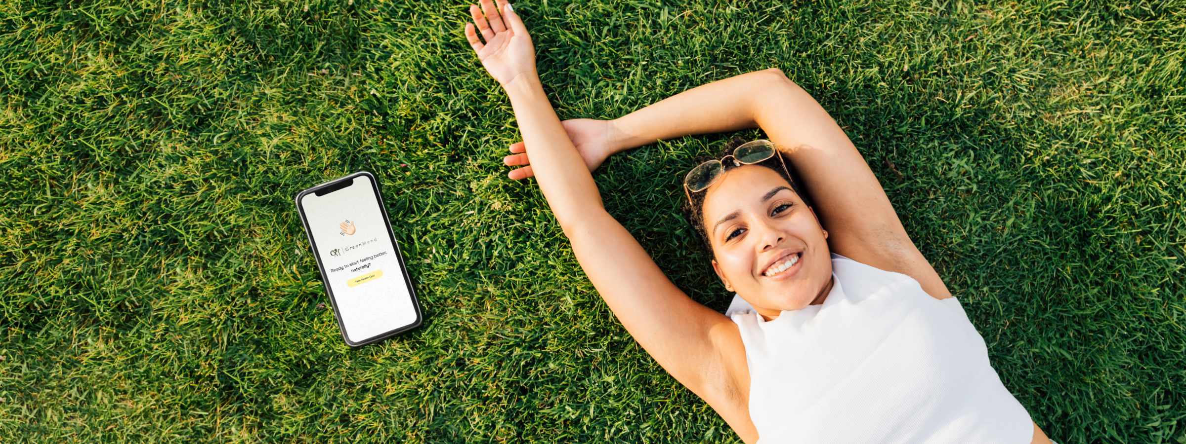A smiling woman lies on grass with her arm raised, next to a smartphone displaying a weather app, showing sunny conditions.
