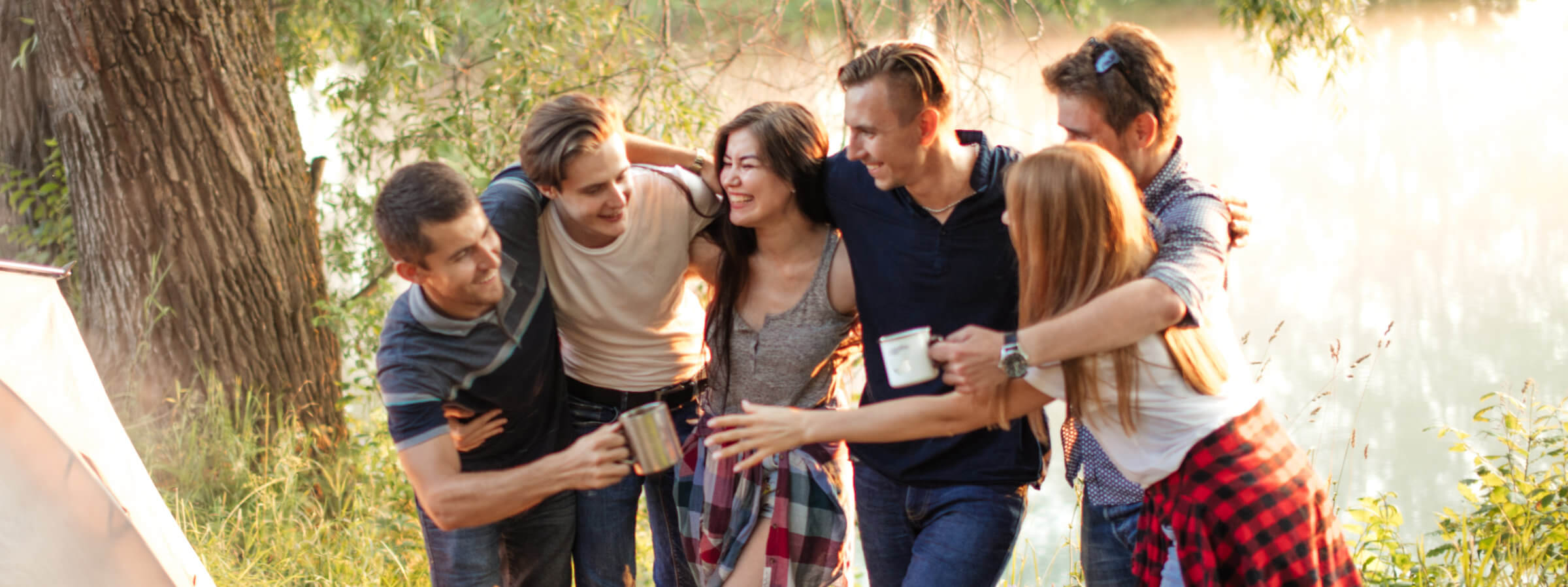 A group of six people stand outdoors near a tree, smiling and holding mugs. Some have their arms around each other. It appears to be a sunny day.