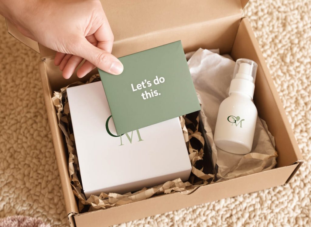 A hand holds a green card reading "Let's do this" over a cardboard box containing paper filler, a product box, and a small pump bottle, both with "GM" branding.