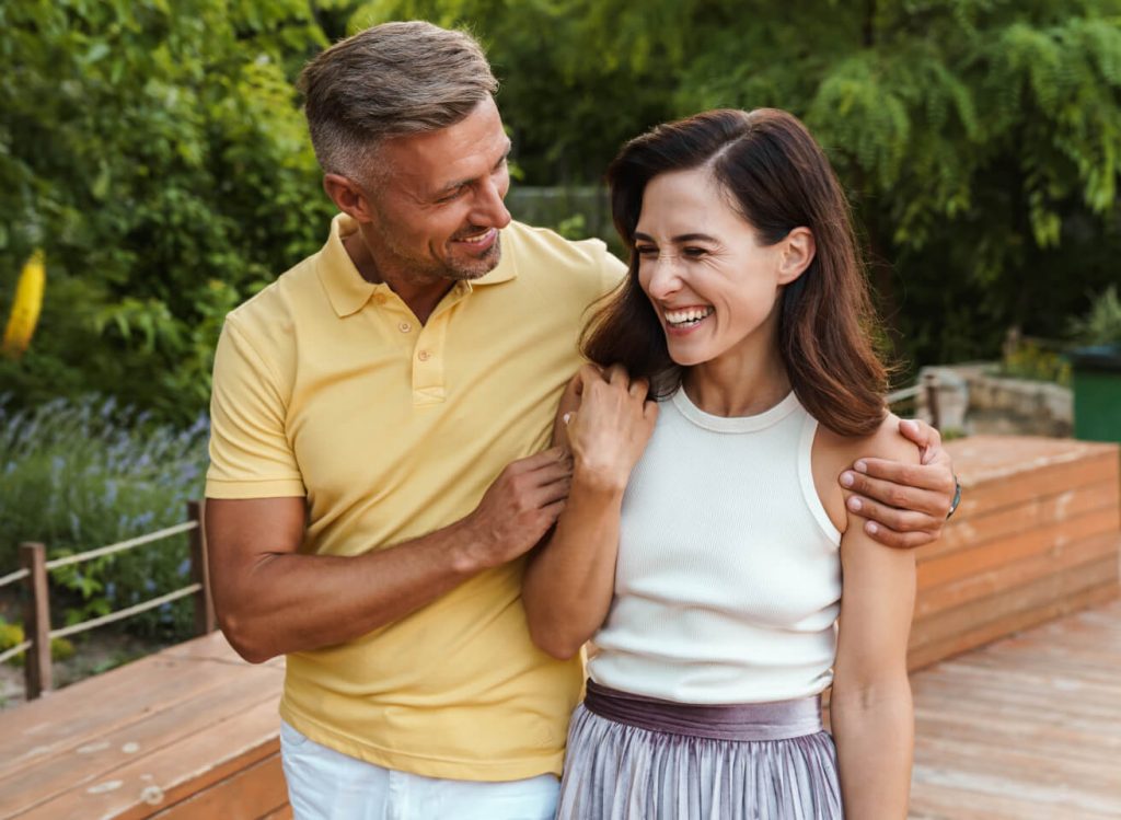 A man and a woman smiling at each other outdoors, standing on a wooden deck surrounded by greenery. The man is wearing a yellow polo shirt, and the woman is in a white top and purple skirt.