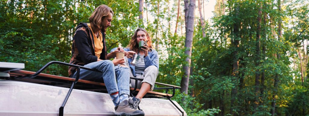 Two people sitting on a vehicle roof in a forest, sharing a meal and drink. The scene is surrounded by lush green trees.