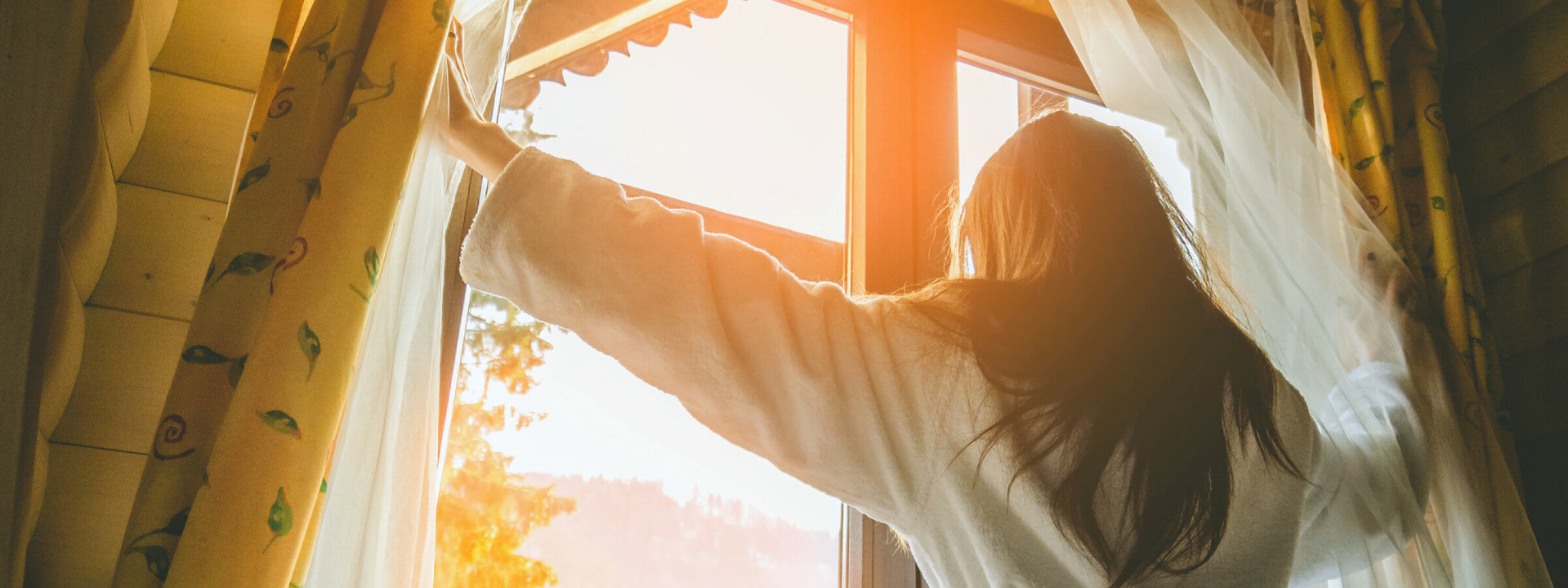 A woman in a robe opens curtains to a bright, sunny view outside a window, letting light into the room.