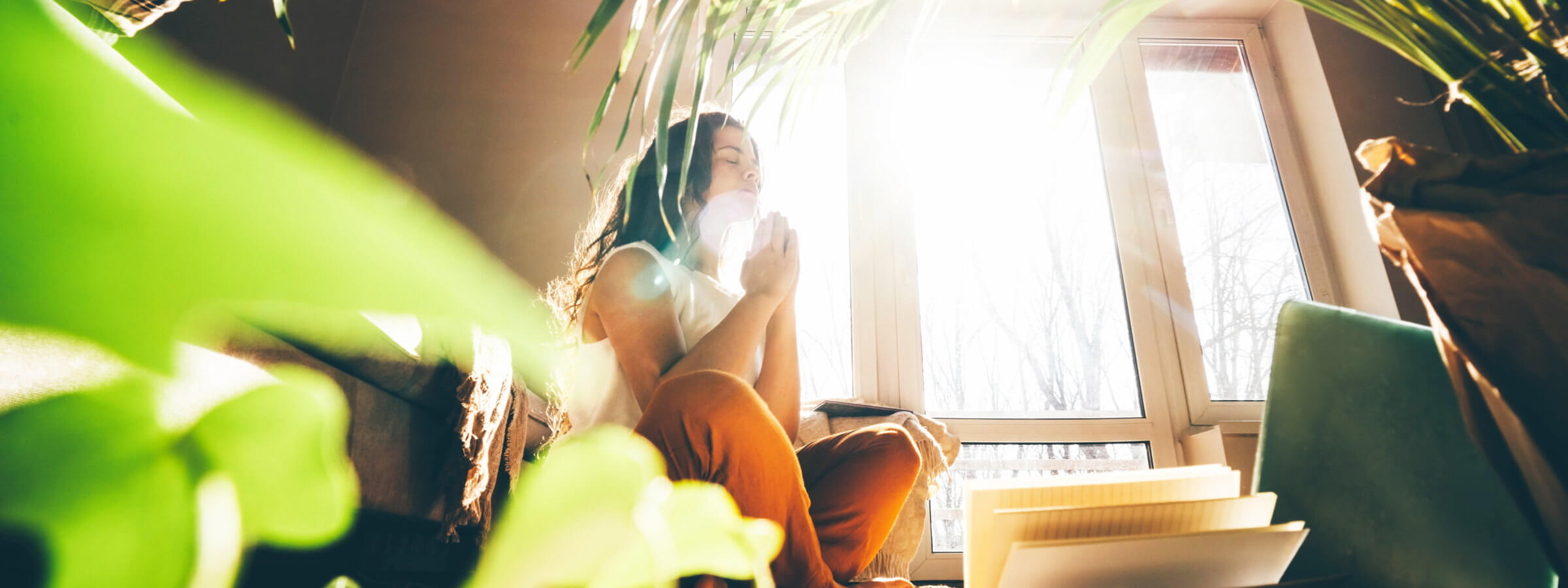 Woman sitting cross-legged in a sunlit room, meditating with closed eyes, surrounded by leafy plants and open notebooks.