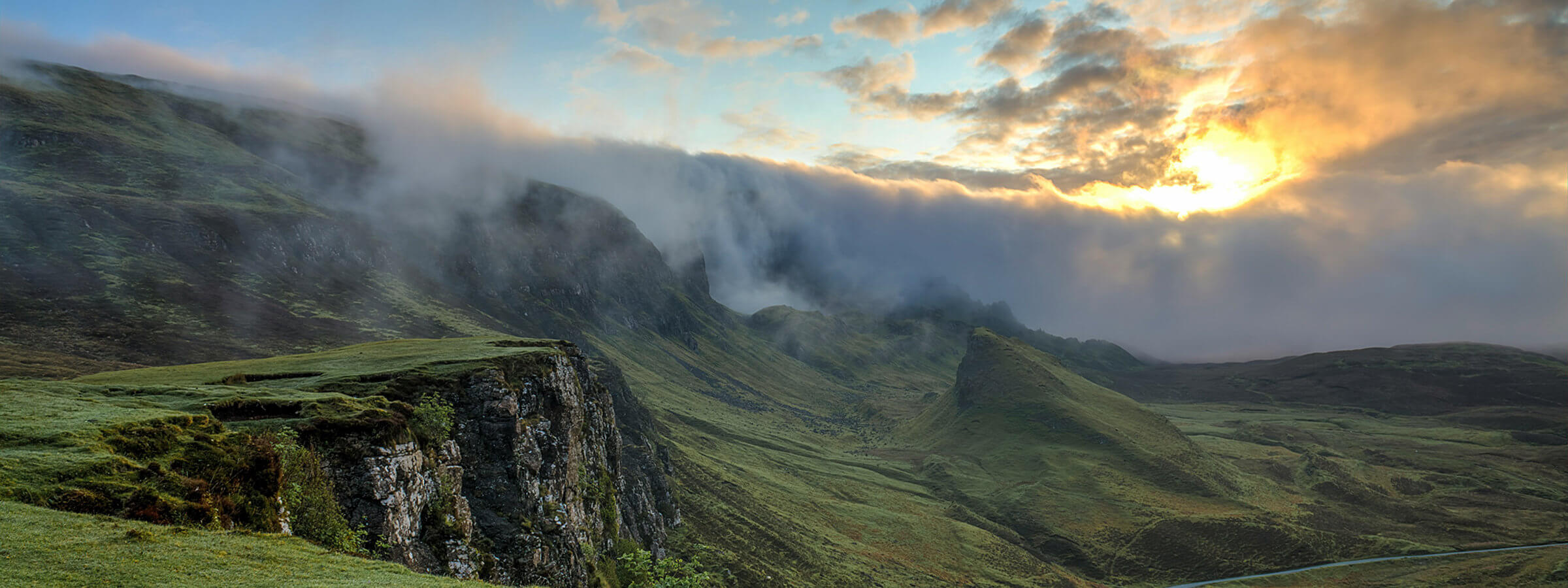 Misty hills and cliffs at sunrise with clouds partially covering the sky, casting a golden light on the landscape.
