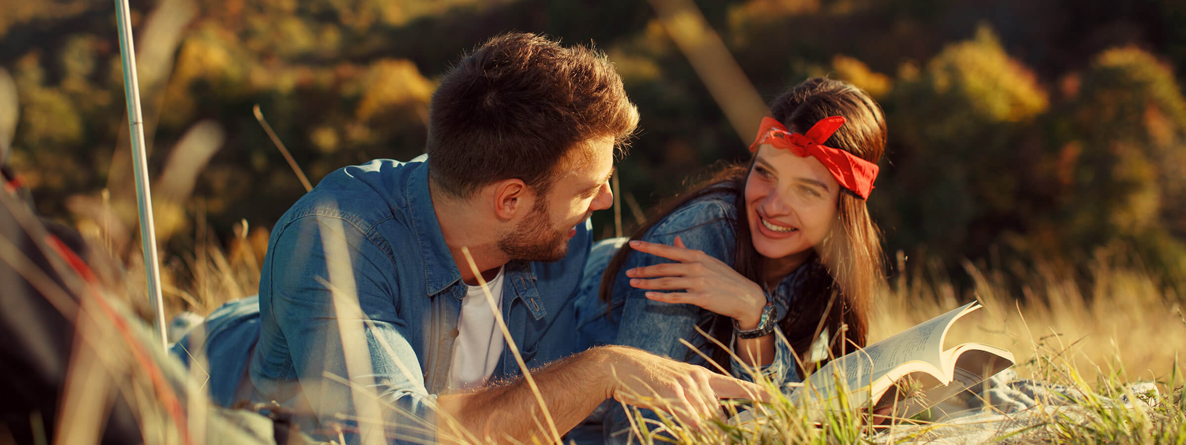 A man and woman lie on grass in a field, smiling and reading a book. The woman wears a red headband. The background shows blurred trees.