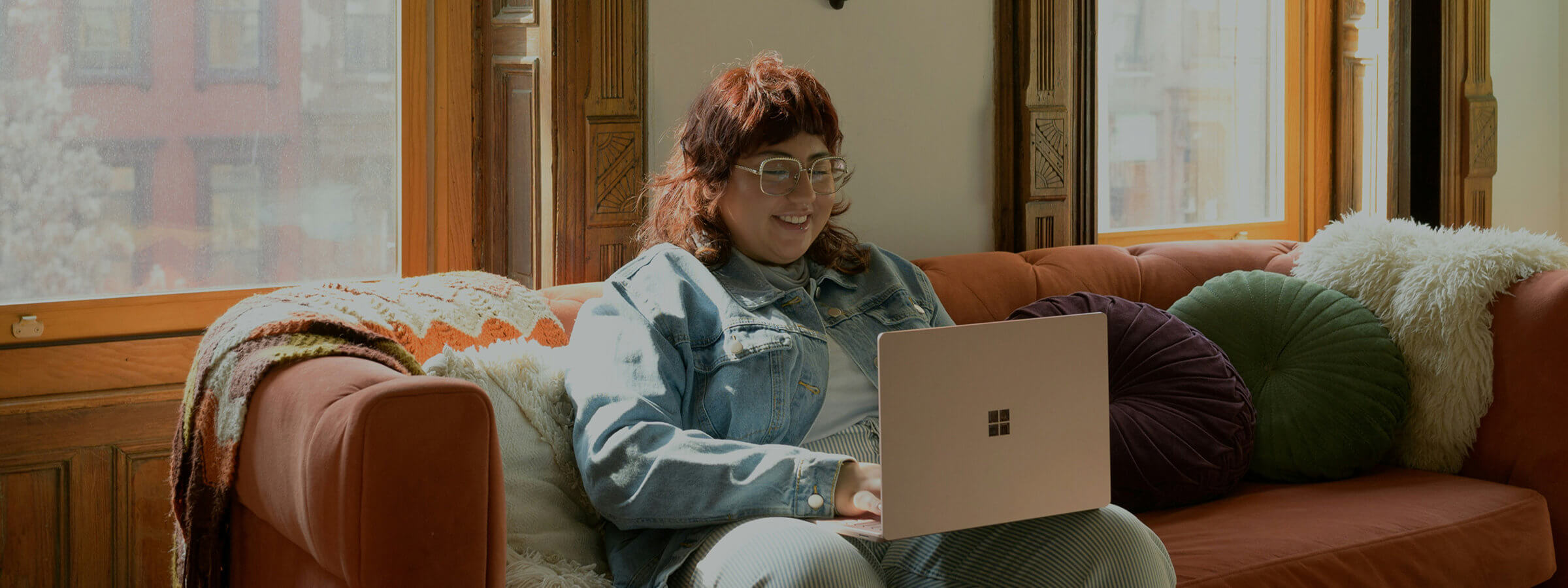A person sitting on a sofa uses a laptop, smiling. Sunlight streams through large windows, and the sofa has colorful cushions and a patterned blanket.