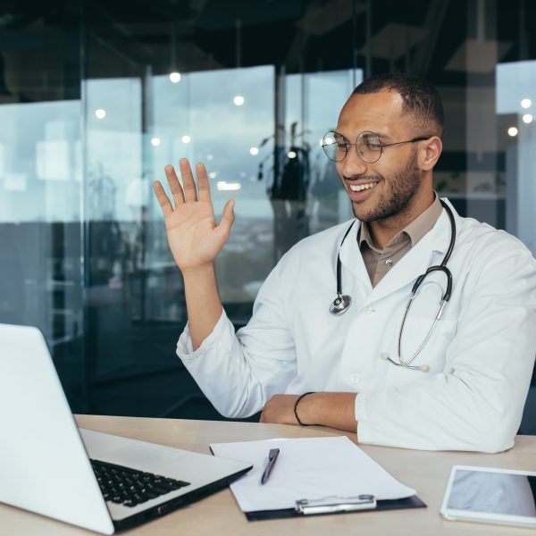 A healthcare provider in a white coat and stethoscope waves while sitting at a desk with a laptop, tablet, and notepad, ready for informed health conversations in a modern office.