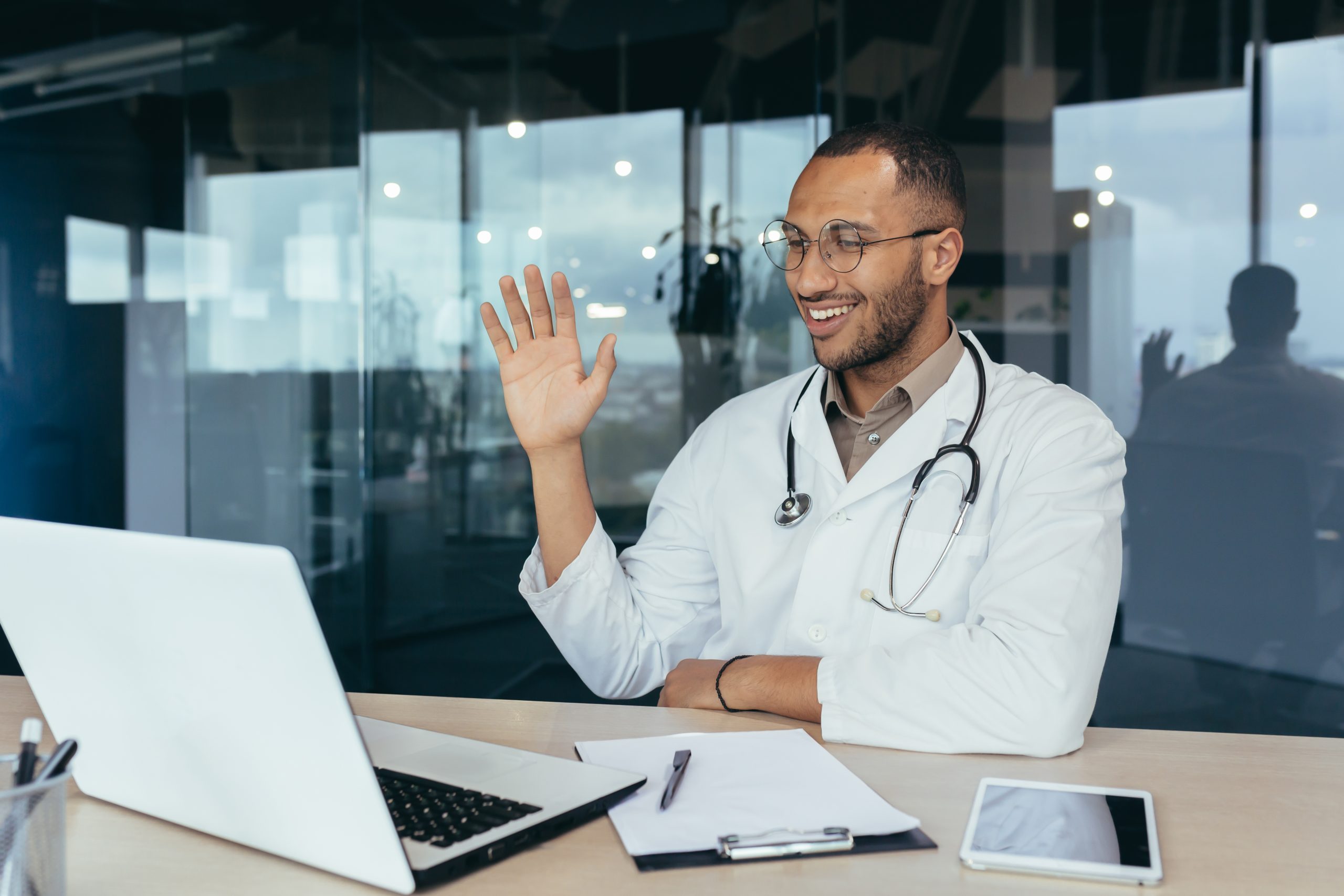 A healthcare provider in a white coat and stethoscope waves while sitting at a desk with a laptop, tablet, and notepad, ready for informed health conversations in a modern office.