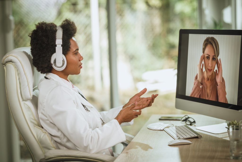 A doctor wearing headphones sits at a desk, engaging in informed discussions during a video call with a patient displayed on the computer screen.