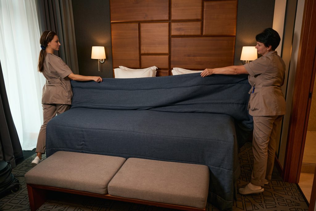 Two housekeepers in uniform efficiently make a bed with a dark blue cover in a hotel room, accented by elegant wooden paneling and warm wall-mounted lamps.